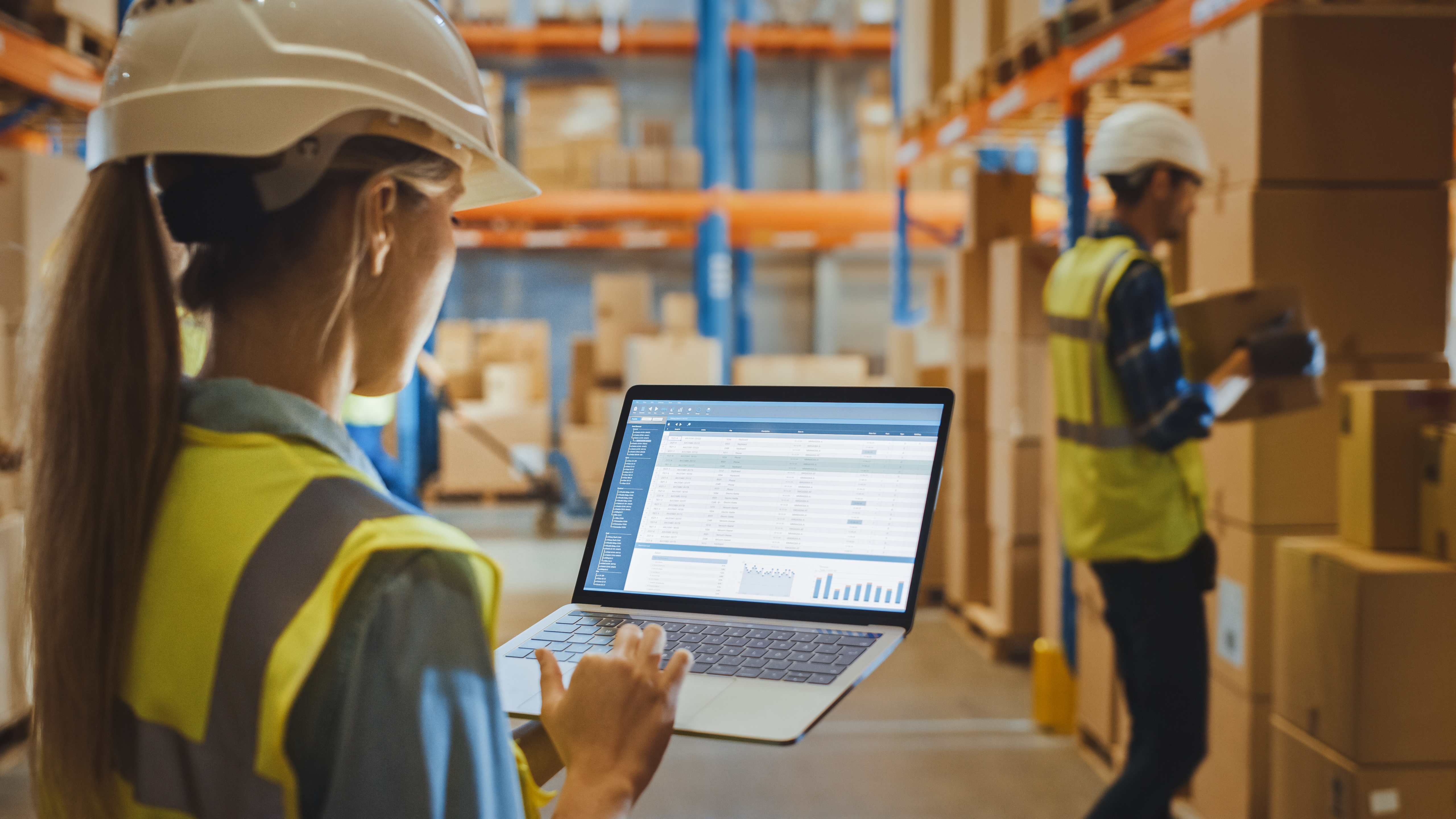 Kvinnelig lagerarbeider med vernehjelm holder en bærbar PC og gjennomgår en rapport om Warehouse Profitability.